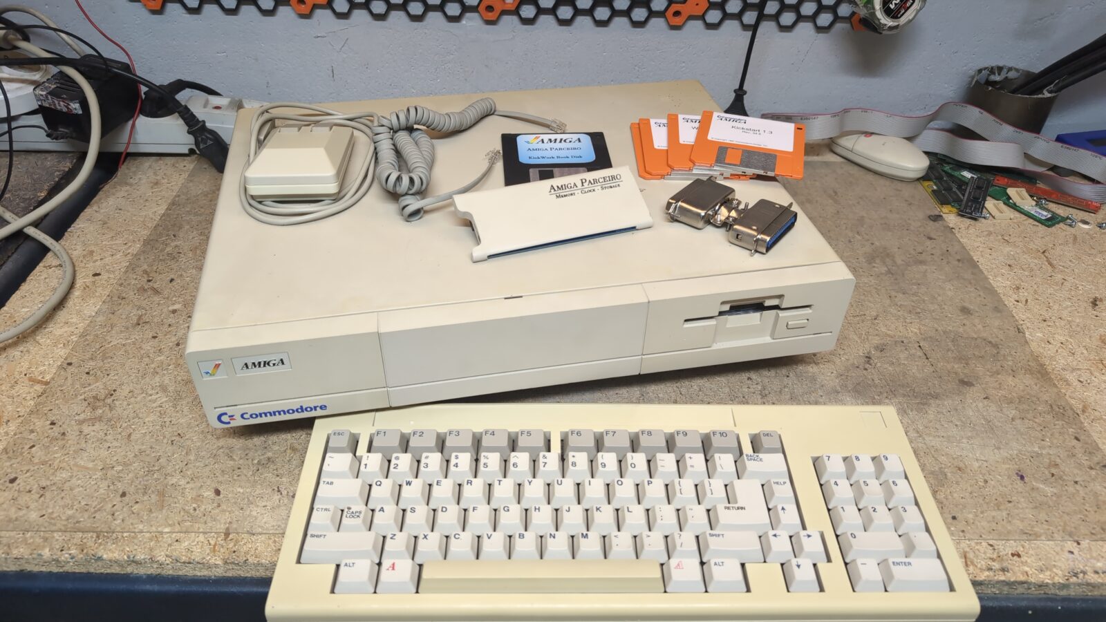 A vintage Commodore Amiga computer with a keyboard, mouse, software disks, a manual, and a connector on a workbench Tools and cables are visible in the background retrohax net