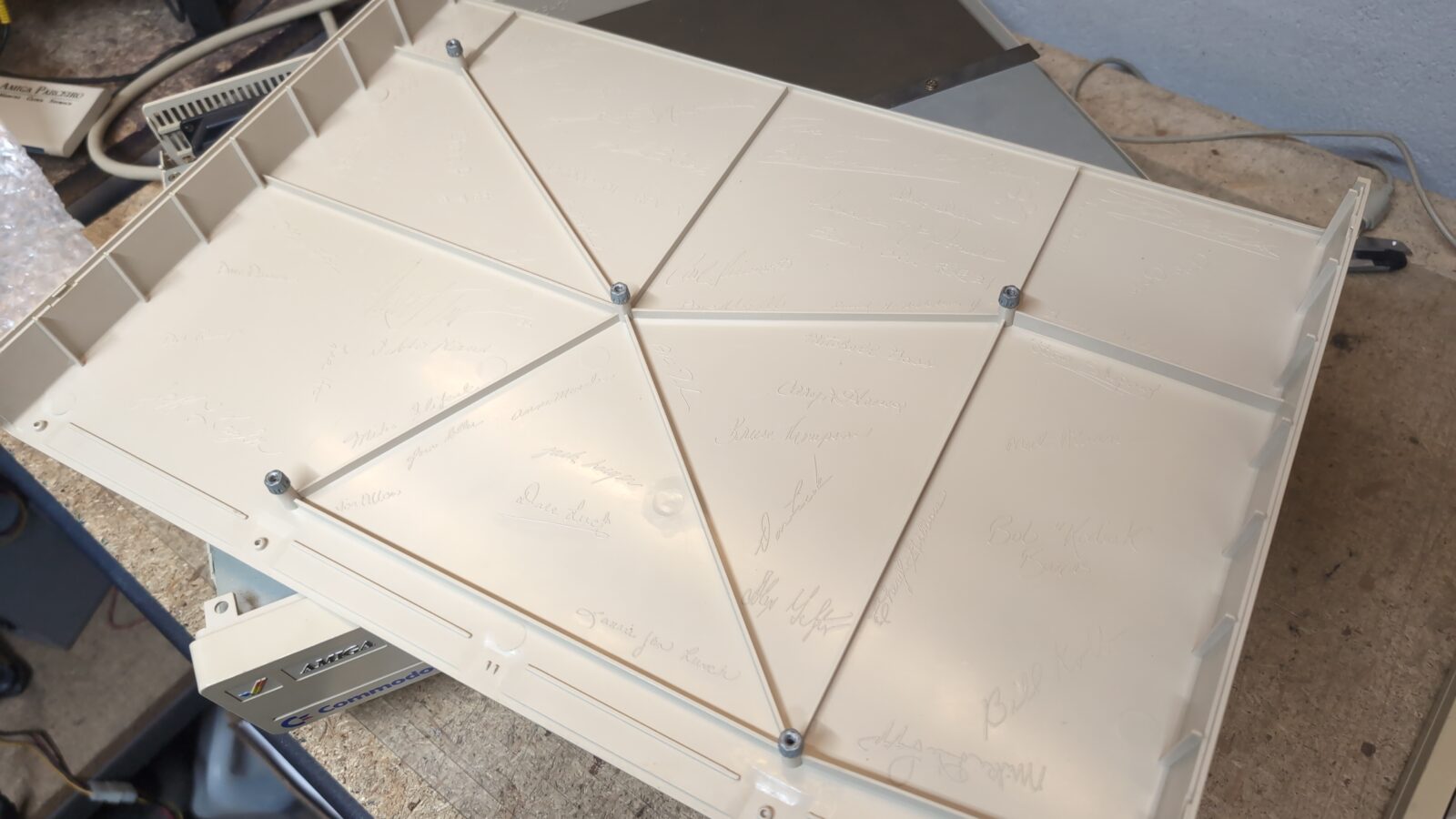 A beige plastic panel with geometric reinforcement lines and several screws, showing numerous faint signatures written across its surface, is placed on a workbench with tools and electronics nearby retrohax net