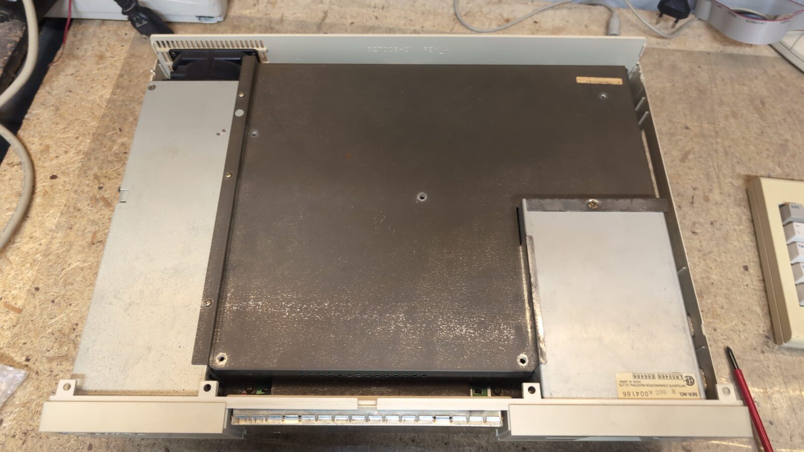 A beige and gray industrial electronic device with a metal cover sits on a workbench; visible are screws, a numeric keypad, and various connectors Cables and tools surround it on the surface retrohax net
