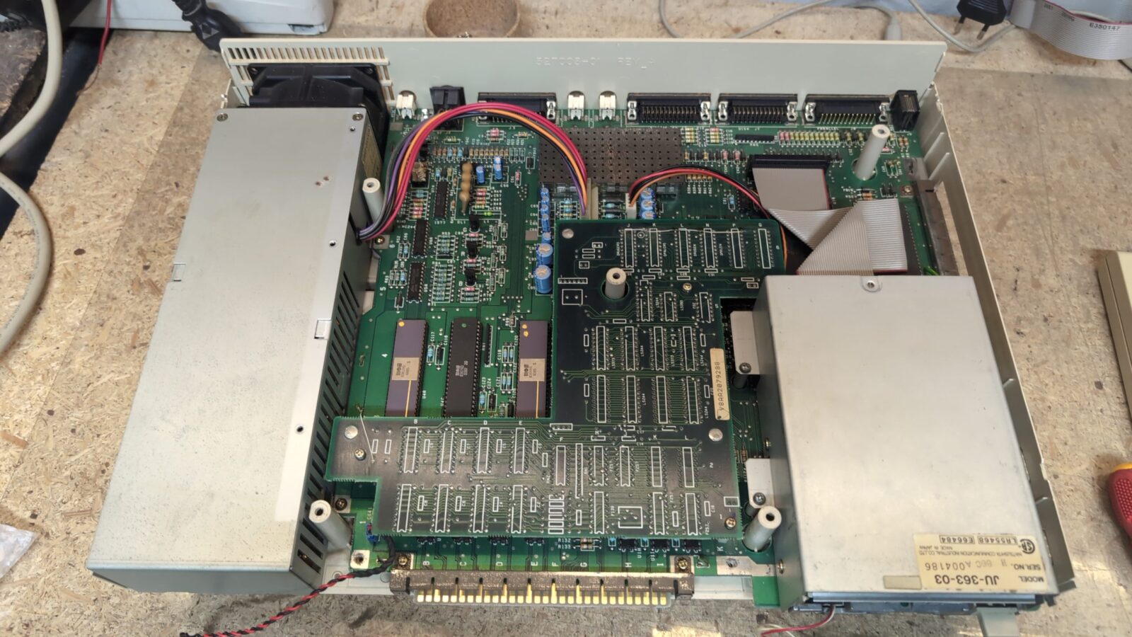 An open electronic device showing a green printed circuit board with various chips, capacitors, connectors, ribbon cables, and two large metal shielded sections, placed on a wooden surface retrohax net