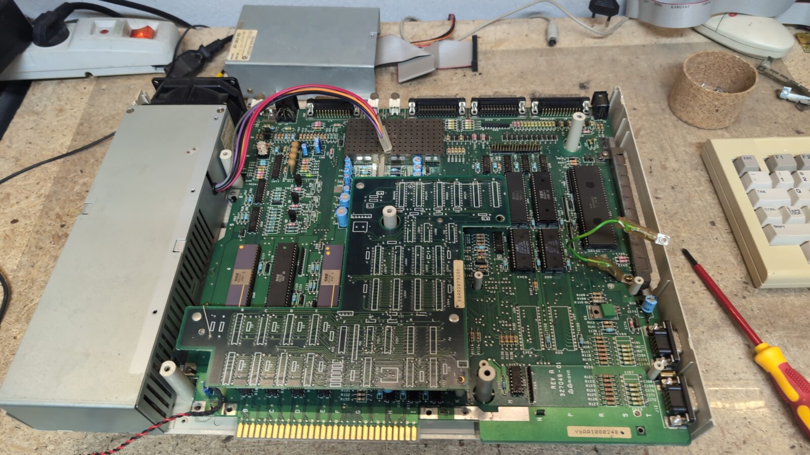 An open vintage computer showing its internal green circuit board, various electronic components, expansion cards, connected wires, and part of a gray keyboard on a workbench with tools and other hardware in the background retrohax net