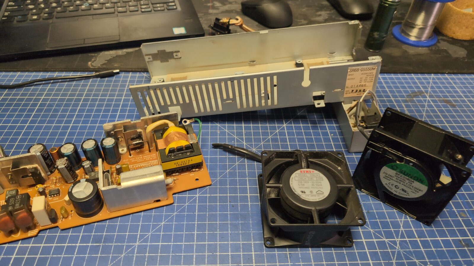 Disassembled electronics on a blue grid mat, including a power supply circuit board, metal casing, two cooling fans, and a partially visible laptop in the background retrohax net