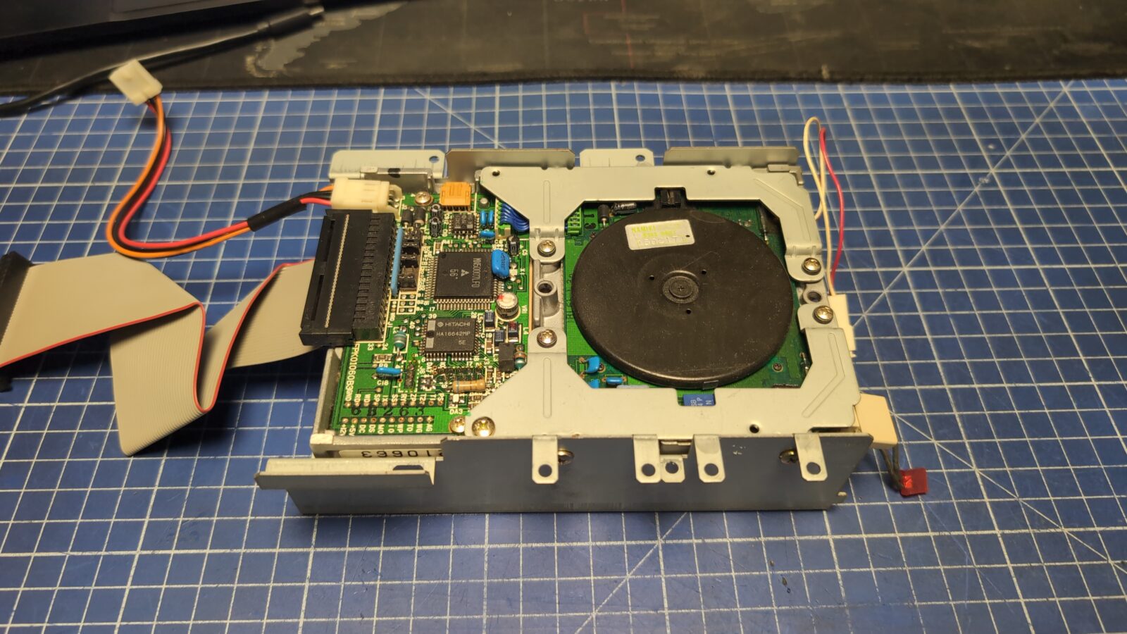 An open floppy disk drive with exposed circuit board and internal components is placed on a blue grid cutting mat, with ribbon cables attached and visible retrohax net