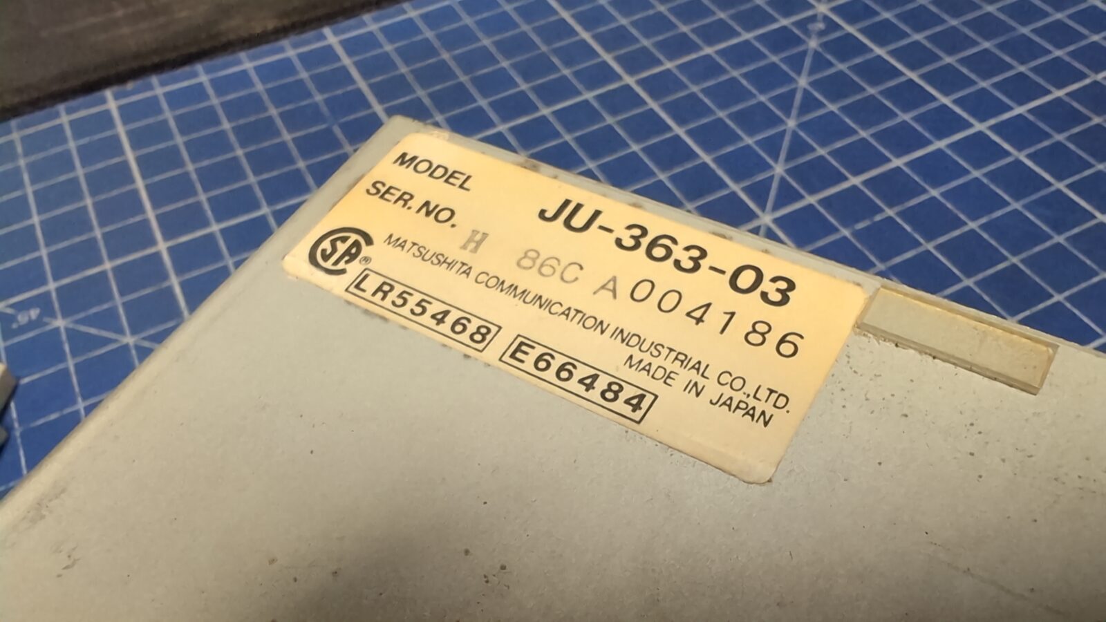 Close up of a label on a gray device showing model JU , serial number CA, and Matsushita Communication Industrial Co , Ltd Made in Japan The label is on a blue grid cutting mat retrohax net