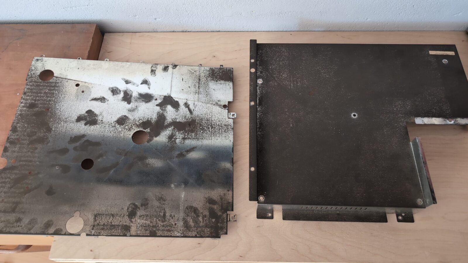 Two rectangular metal panels with circular holes and smudgy black marks lie side by side on a light wooden surface, with one panel positioned above the other retrohax net