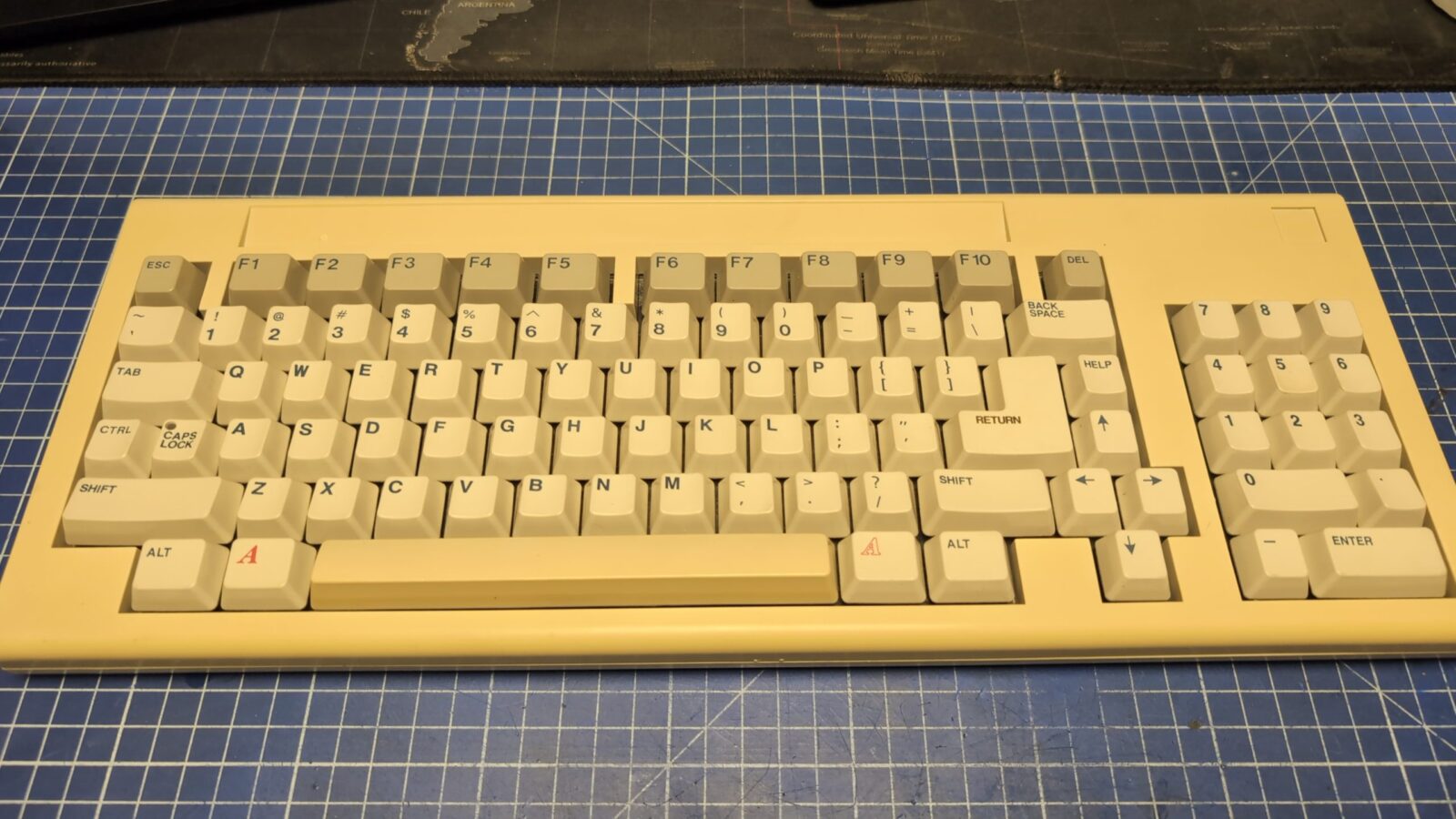 A beige mechanical keyboard with white keycaps and black legends is placed on a blue grid patterned cutting mat The keyboard has a compact layout and appears vintage retrohax net