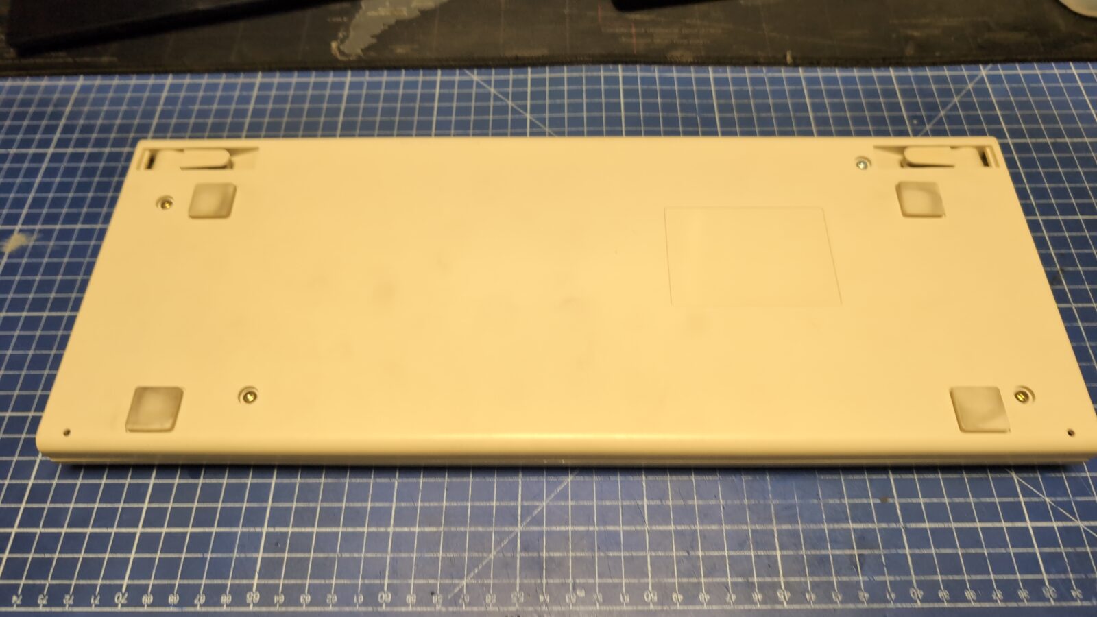 A beige rectangular keyboard case is placed upside down on a blue cutting mat with a white grid pattern The case has two rubber feet, two metal screws, and a small rectangular indentation on its underside retrohax net
