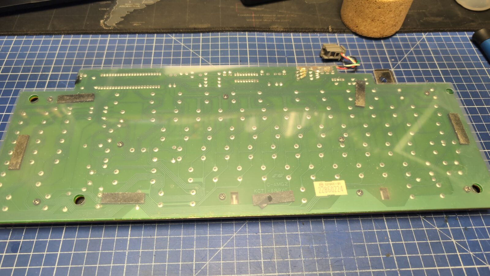 A green circuit board with numerous solder points and electronic components is placed on a blue grid cutting mat Some wires are attached in the top right corner, and a cork object is partially visible in the background retrohax net