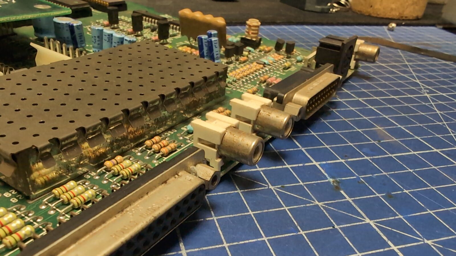 Close up of an electronic circuit board with various components, including resistors, connectors, and ports, resting on a blue grid cutting mat retrohax net