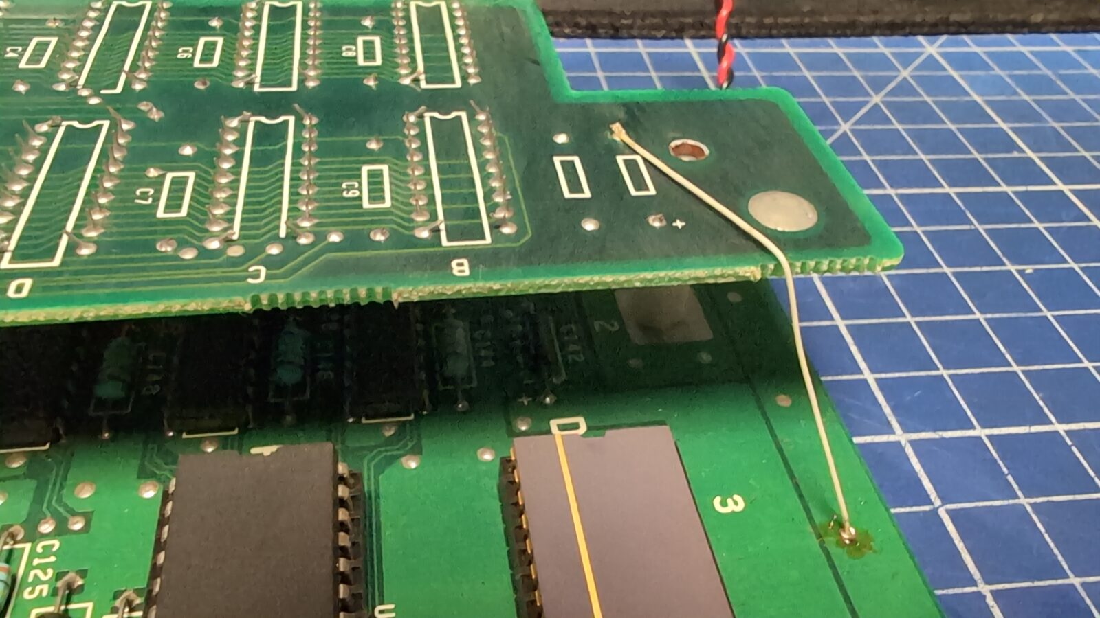 A close up of two stacked green circuit boards, connected by a thin yellow wire, resting on a blue grid patterned work mat Electronic components and traces are visible on the boards retrohax net