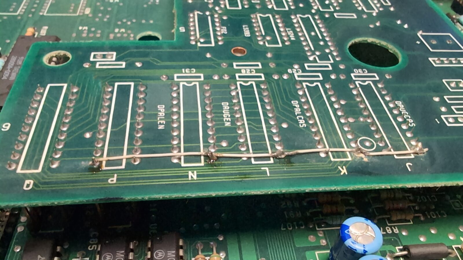 Close up of a green printed circuit board (PCB) with visible soldered connections, labeled sections, and electronic components, including a blue cylindrical capacitor in the lower right corner retrohax net